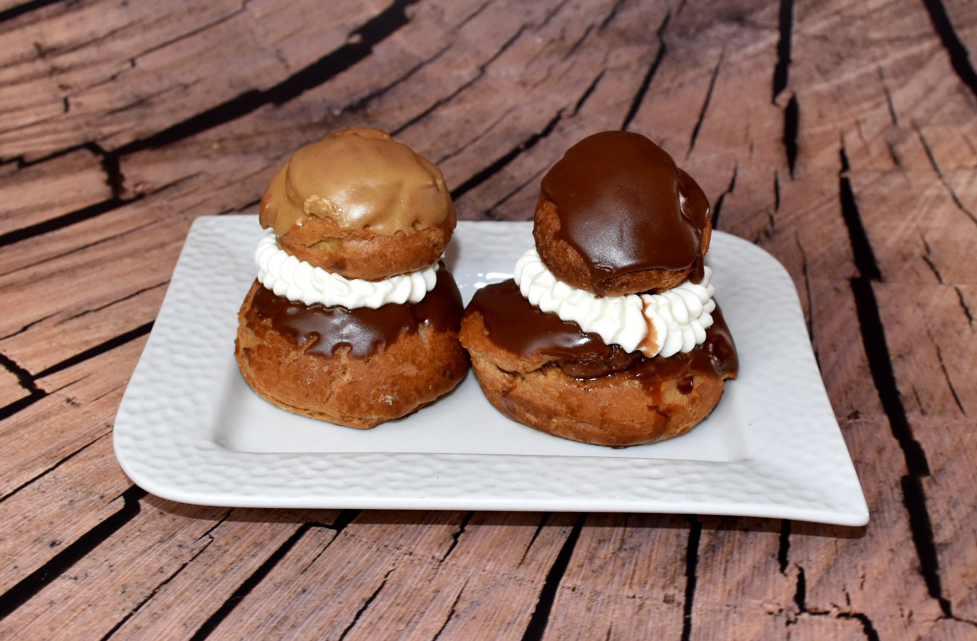 Deux choux garnis, nappés de chocolat, sur une assiette blanche, posés sur une table en bois. Religieuse