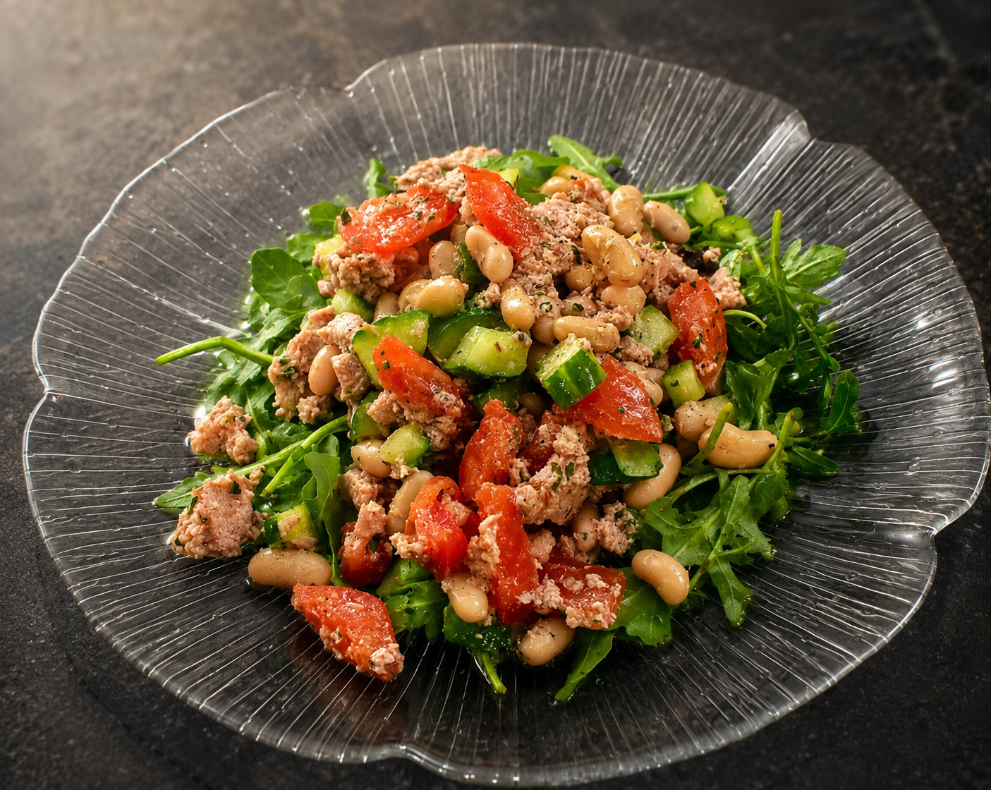Salade dans une assiette transparente : roquette, concombre, tomates, thon émietté et haricots, sur fond sombre.