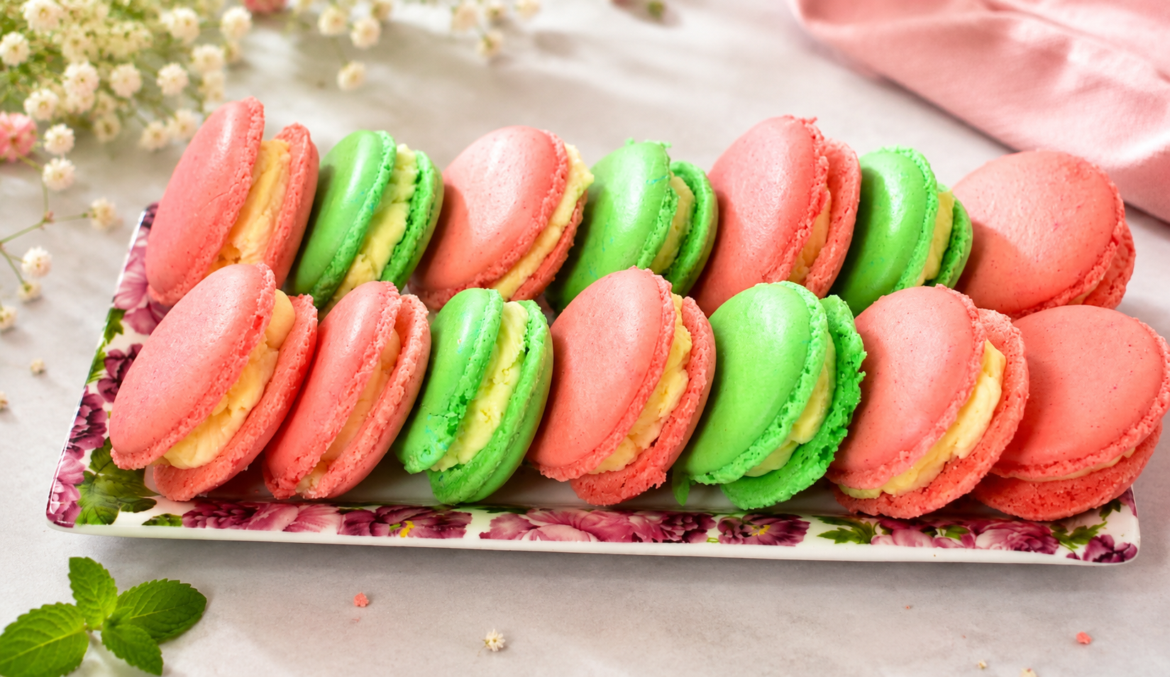Assortiment de macarons roses et verts fourrés, disposés sur un plateau fleuri, décorations printanières.