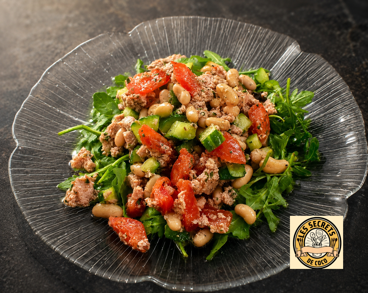 Salade dans une assiette transparente : roquette, concombre, tomates, thon émietté et haricots, sur fond sombre.