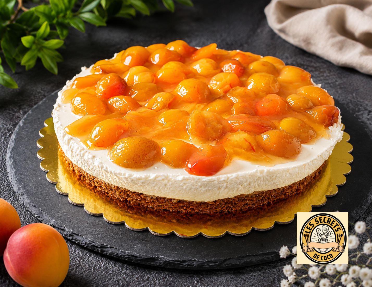 Gâteau au fromage nappé de mirabelles brillantes, sur assiette dorée, fond sombre avec feuilles vertes et tissu.