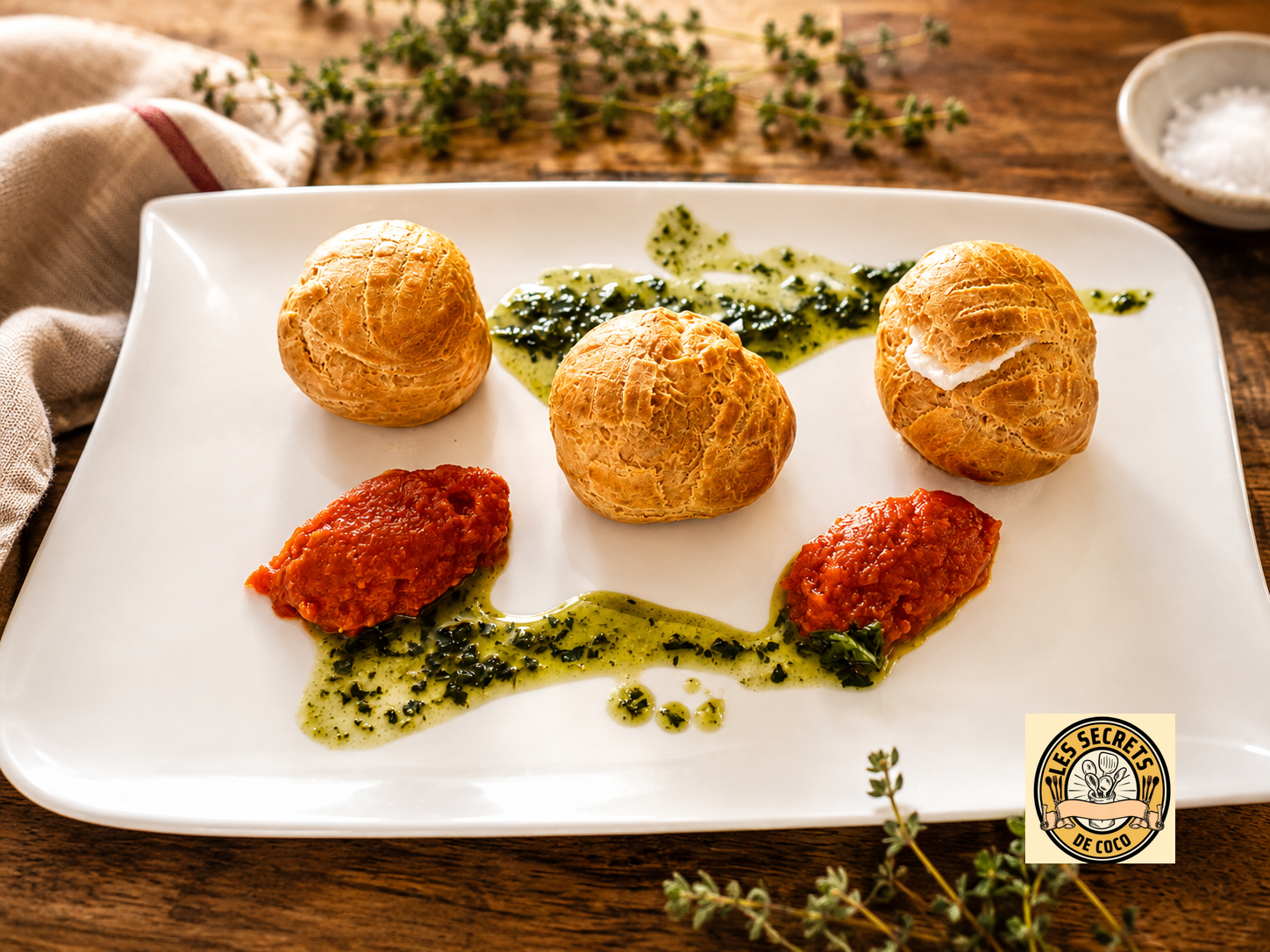 Trois choux garnis sur assiette blanche, sauce verte et deux morceaux rouges, table rustique.