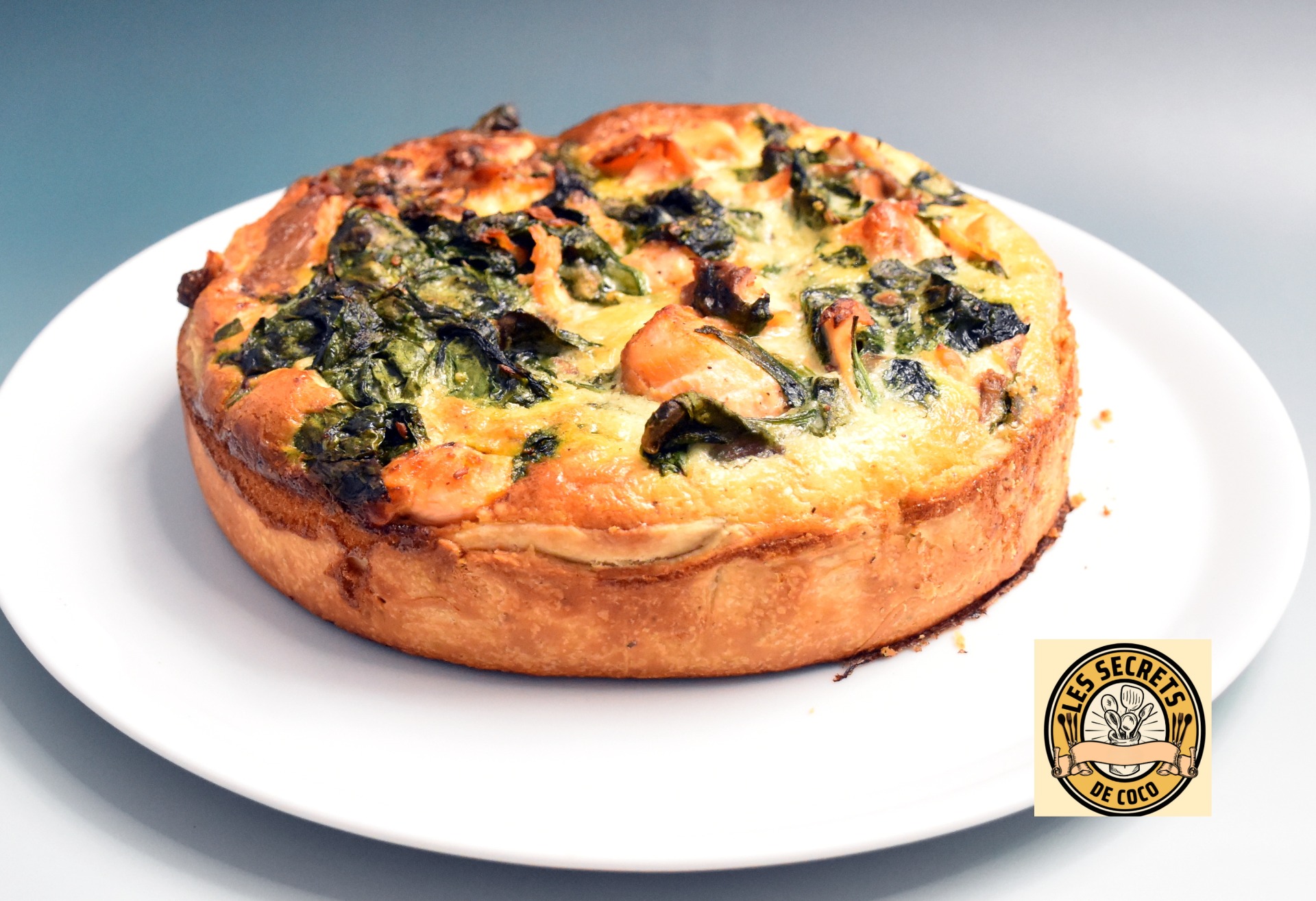 Tarte salée ronde aux épinards et saumon, tranches visibles, sur assiette blanche, fond bleu clair.