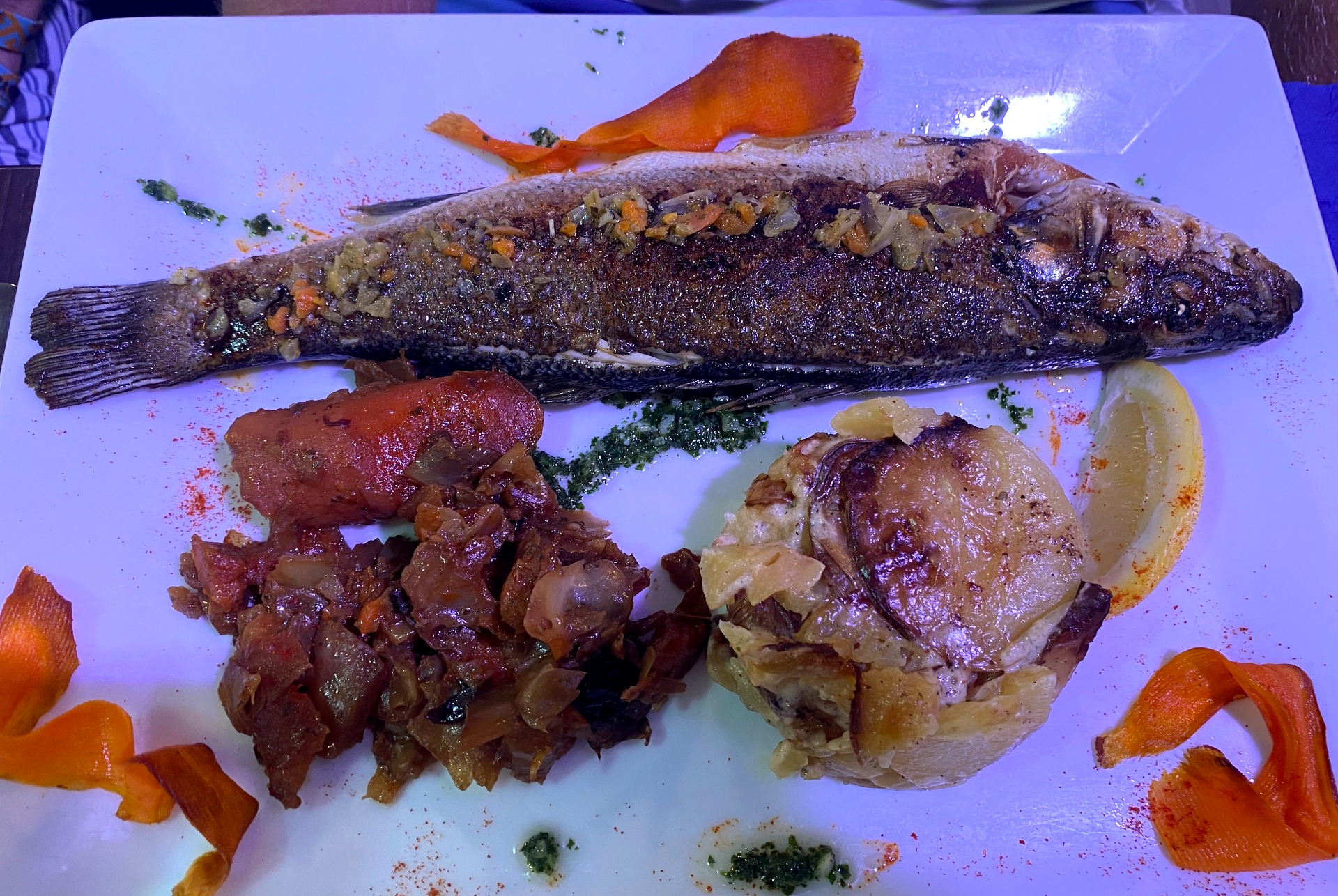 Poisson grillé (bar) sur assiette blanche, couvert d’herbes et d’épices, avec carottes et garniture hachée.