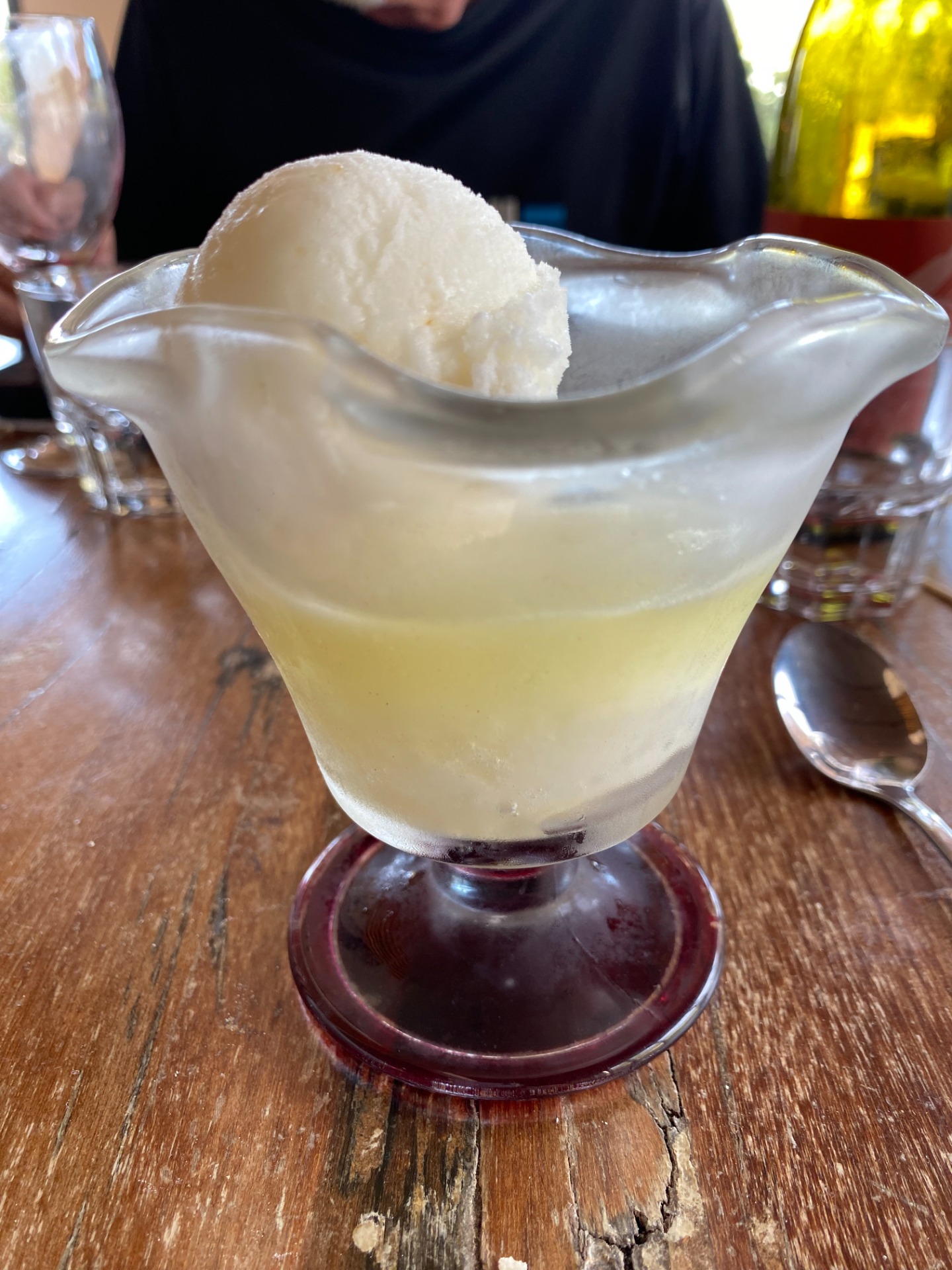 Verre à cocktail jaune pâle avec glaçon, posé sur une table en bois, autres verres flous en arrière-plan. sorbet arrosé limoncello citron