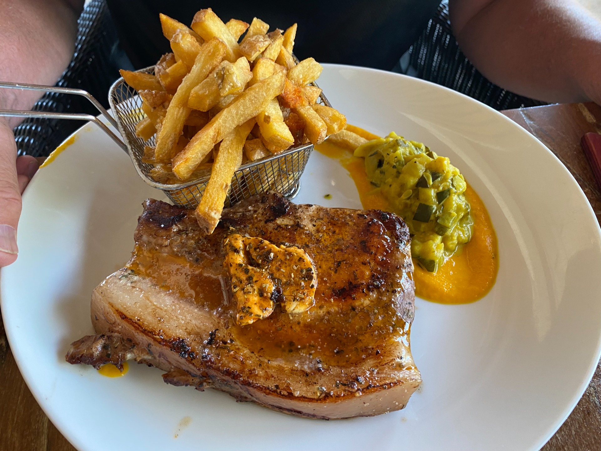 Assiette avec côte de porc grillée, frites en panier, purée verte et sauce jaune, vue rapprochée.
