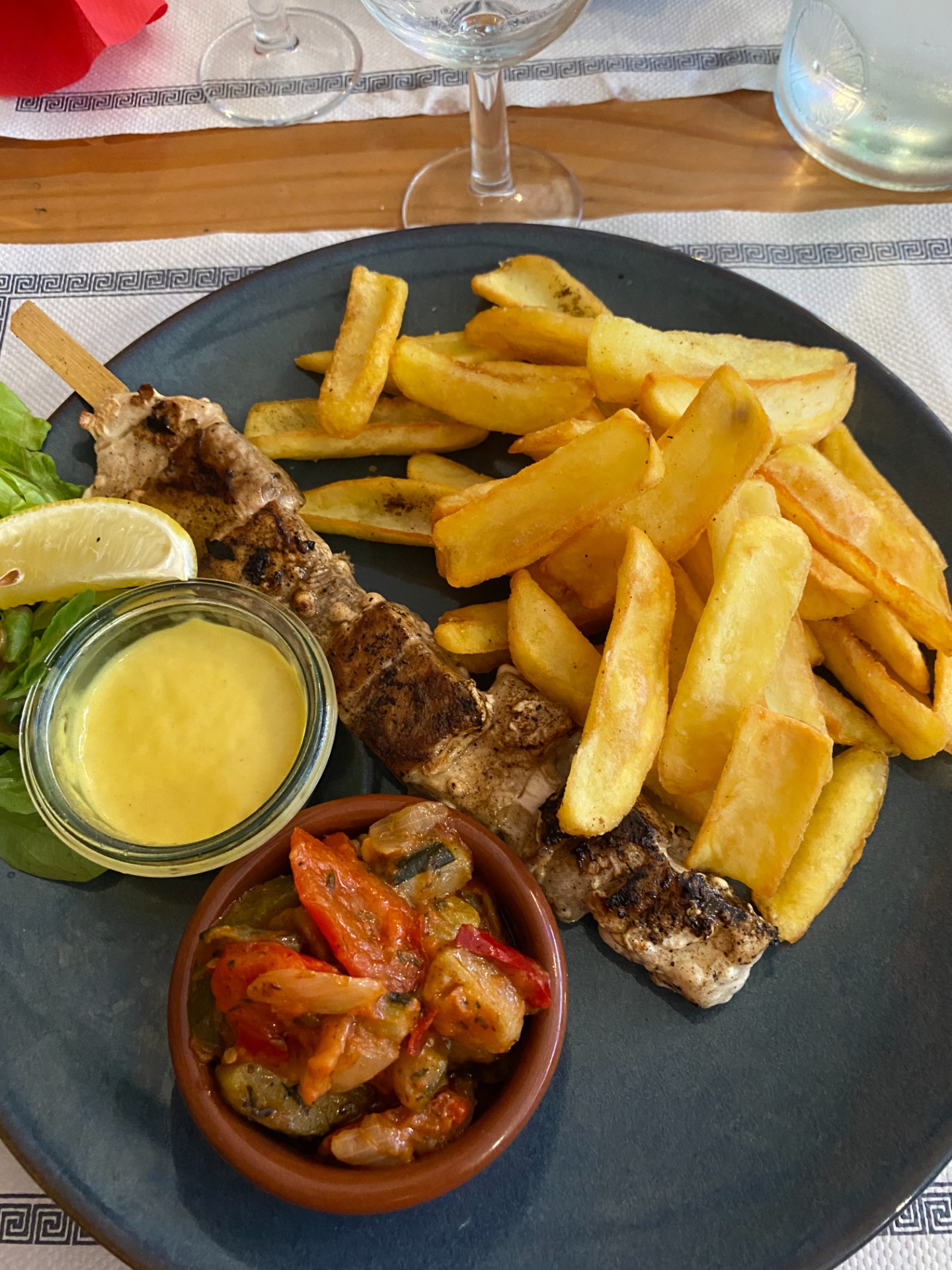 Assiette noire avec frites dorées, morceaux de poisson grillé, salade verte, citron et sauce en pot.