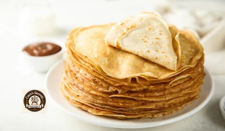 Pile de crêpes dorées empilées sur une assiette, une crêpe pliée au-dessus, fond clair.